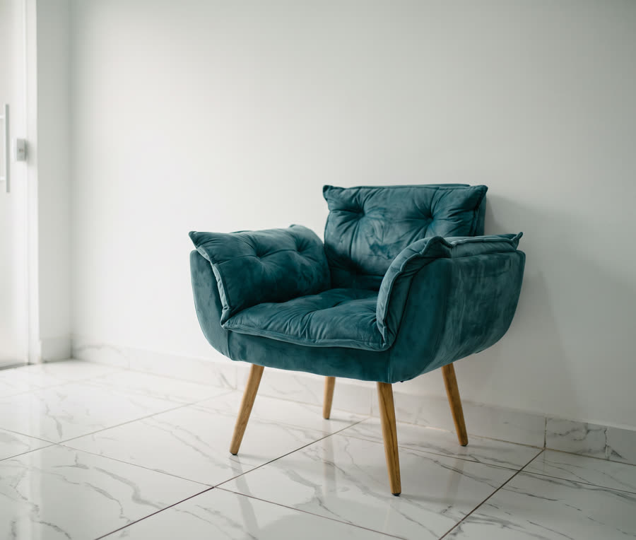 Poltrona de veludo azul-petróleo com pés de madeira na sala de espera, iluminada pela luz lateral